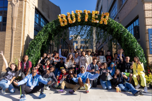 Image: A group of smiling young people pose at the entrance of the BUTTER Art Fair, beneath a floral archway. BUTTER is spelled out in yellow flowers at the top of the arch. Photograph by Simone Wallis, image courtesy of GANGGANG.